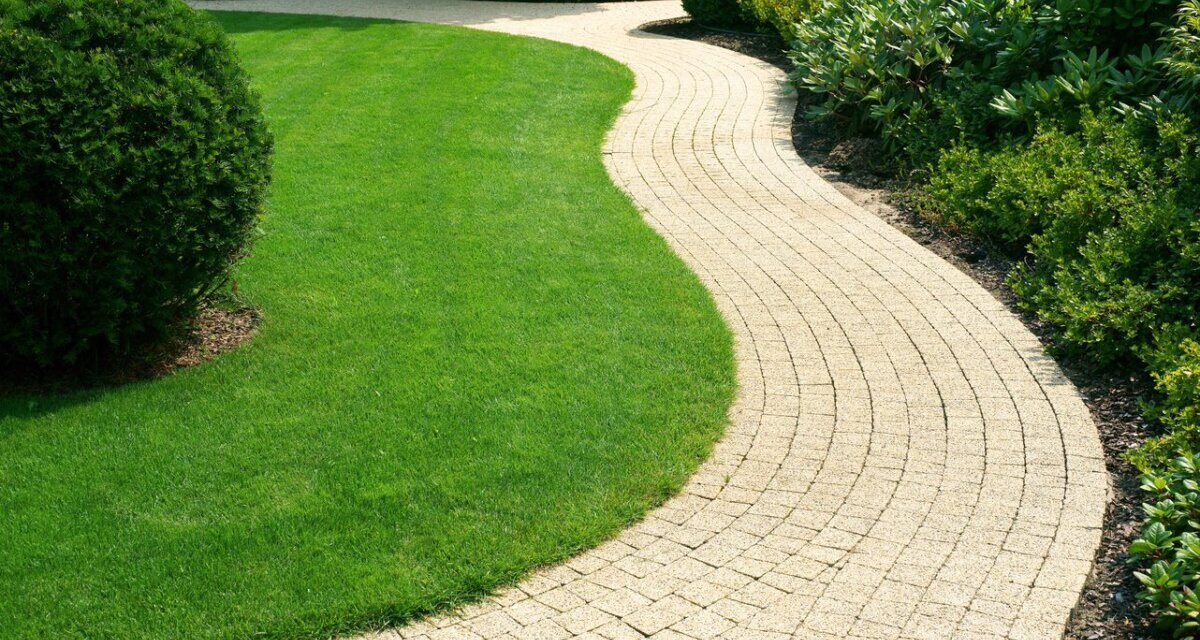 Zadbany ogród o nieregularnych kształtach ze ścieżką w kształcie fali, ułożoną z betonowej kostki oraz z pięknie przyciętym, intensywnie zielonym trawnikiem.